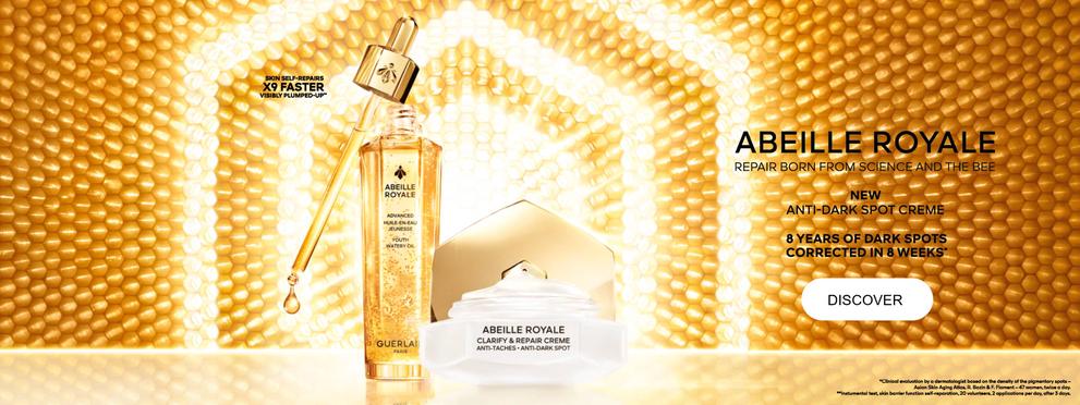  The image is an advertisement for Guerlain's Abeille Royale line featuring a serum and a cream, set against a golden background of leaves partially dusted with white, interwoven with a sparkling green ribbon with the slogan "Live in Fantasy".